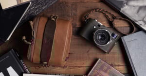 A vintage-style camera with a braided strap and a brown leather case sit on a wooden table surrounded by books and magazines.