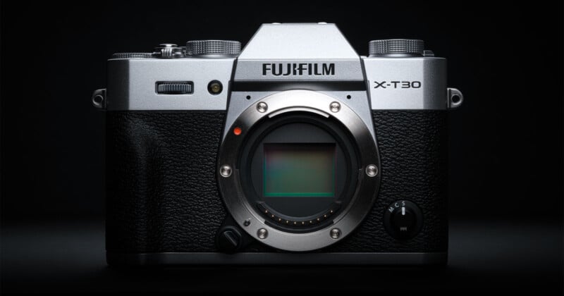 A Fujifilm X-T30 mirrorless digital camera with a silver and black body is shown without a lens, revealing the camera sensor, against a dark background.