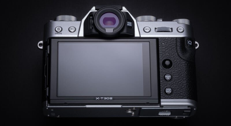 A close-up of the back of a silver digital camera with a large LCD screen, viewfinder, control dials, and several buttons on a dark background.