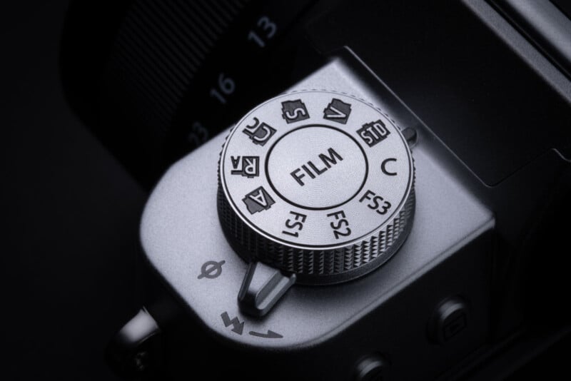 Close-up of a silver camera mode dial labeled "FILM" at the center, surrounded by various symbols and settings. The dial and part of the camera body are visible against a dark background.