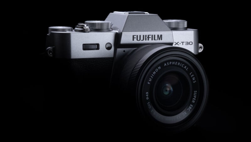 A Fujifilm X-T30 mirrorless digital camera with a Fujinon aspherical lens, shown in a partial side view against a black background. The camera body is silver and black with visible dials and textured grip.