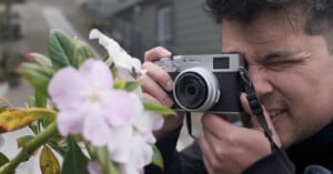 A person holds a camera up to their face, focusing and preparing to take a photo of light pink flowers on a leafy green plant outdoors.