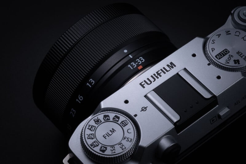 Close-up of a silver Fujifilm camera with a 13-33mm lens, showing detailed dials, buttons, and the "FILM" mode dial, set against a dark background.