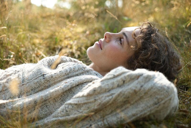 A person with curly hair lies in tall grass, wearing a chunky knit sweater, looking relaxed and gazing upward, surrounded by soft, warm sunlight.