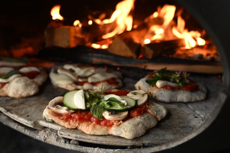 Mini pizzas with fresh toppings, including basil, zucchini, and mozzarella, baking on a stone inside a wood-fired oven with flames and burning logs in the background.