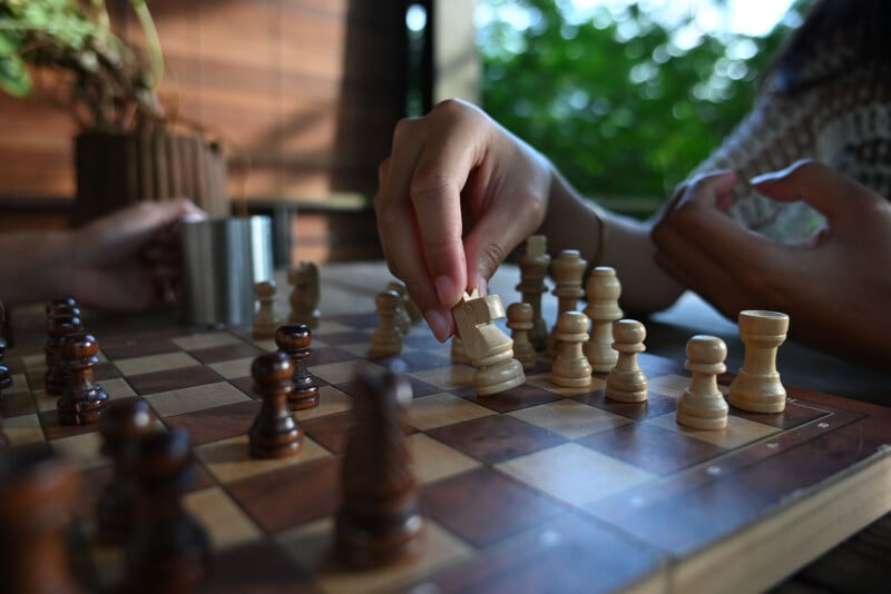 A close-up of two people playing chess outdoors, with one person’s hand moving a white rook on a wooden chessboard. The background shows greenery and sunlight filtering through.