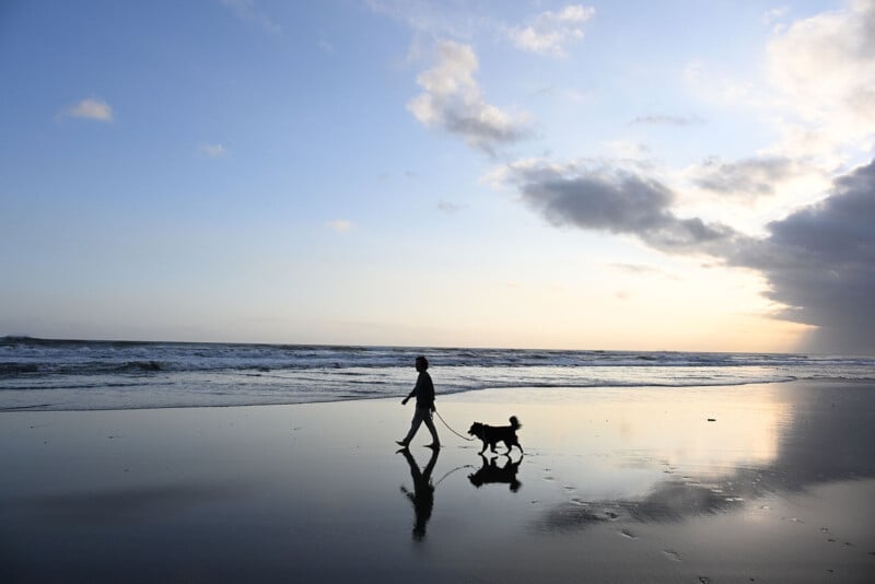A child walks a small dog along the beach at sunset, their reflections visible in the wet sand. The sky is partly cloudy, and gentle waves roll onto the shore.