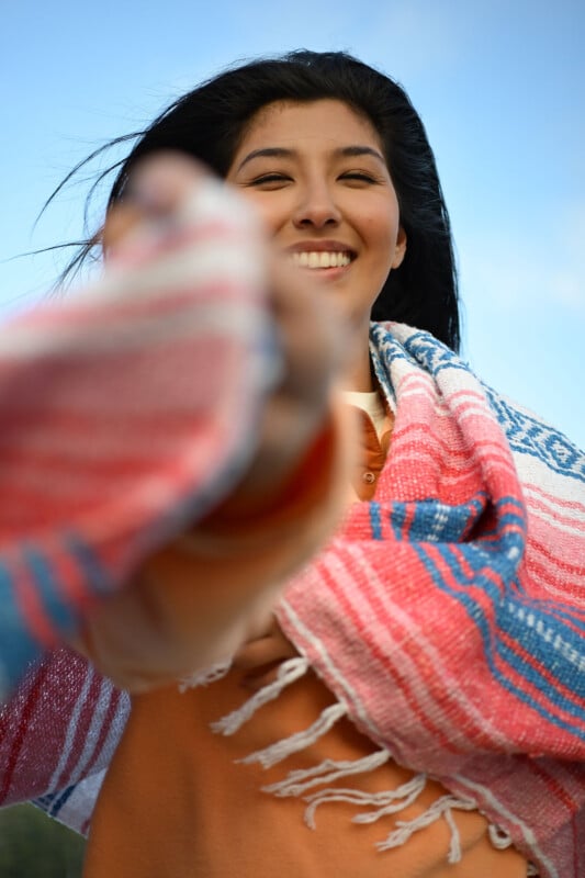 A smiling woman wrapped in a colorful striped blanket holds out her hand toward the camera against a blue sky background.