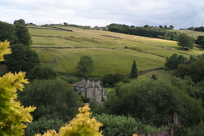 A stone house surrounded by trees sits in a green, hilly countryside under a cloudy sky, with fields and stone walls in the background. Shrubs and flowering plants frame the foreground.