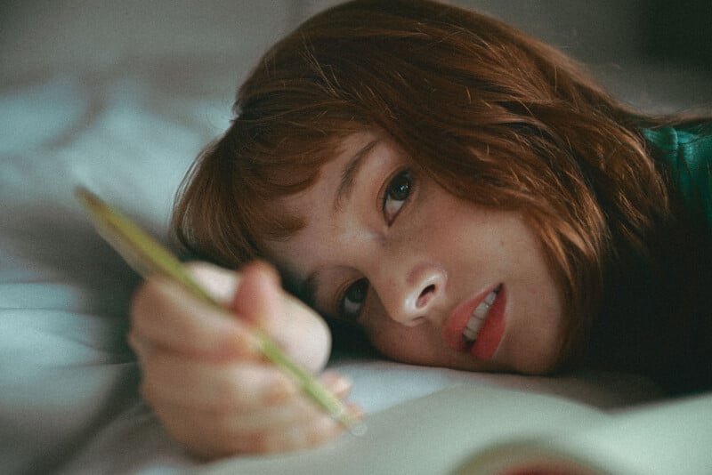 Un joven de largo cabello castaño está acostado en la cama, mirando pensativamente a la cámara y escribiendo en un cuaderno con un lápiz. La iluminación suave crea un ambiente tranquilo e introspectivo.