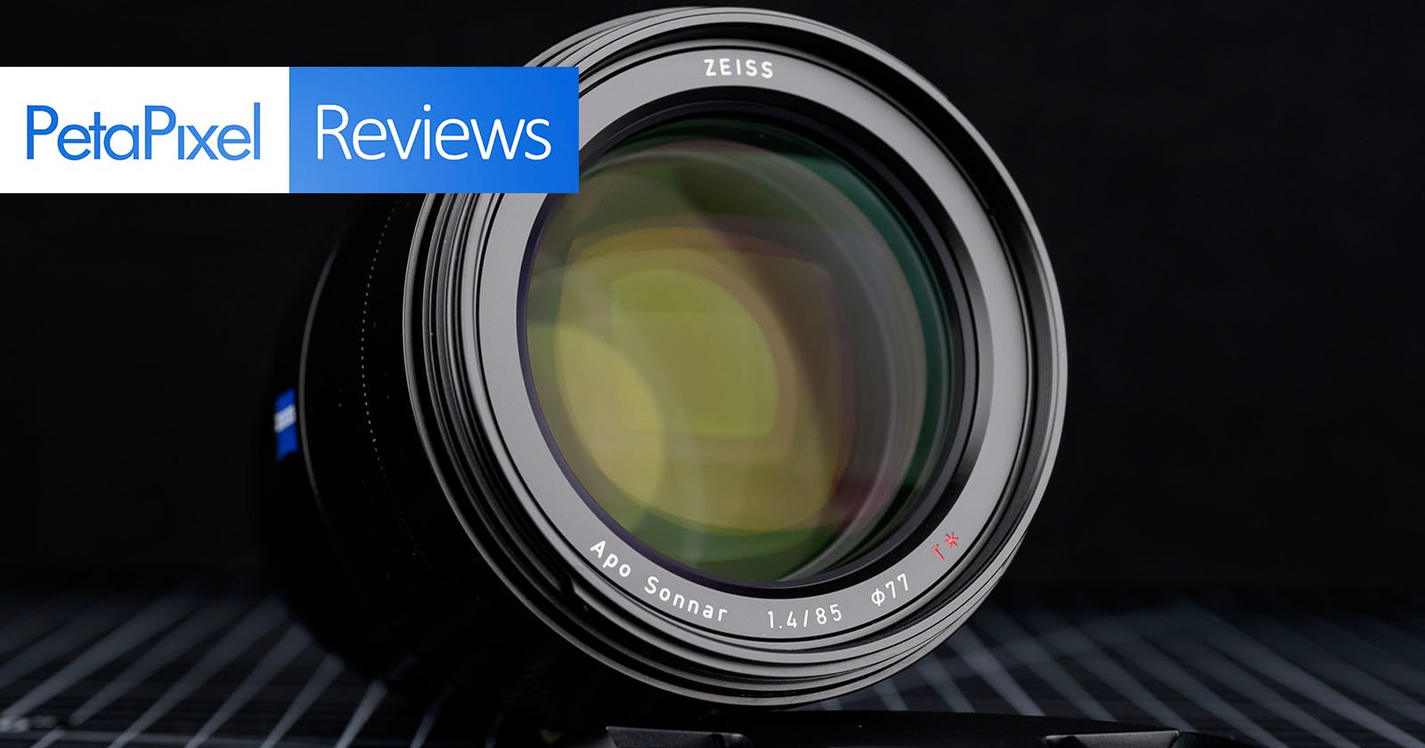 Close-up of a Zeiss Apo Sonnar 85mm f/1.4 camera lens with "PetaPixel Reviews" text overlay in the top left corner, set against a dark background.