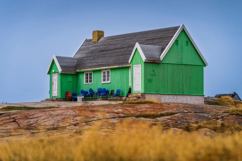 A bright green wooden house with a gray roof sits on rocky terrain, surrounded by yellowish grass. Blue chairs are grouped outside near the house under a clear, blue sky.