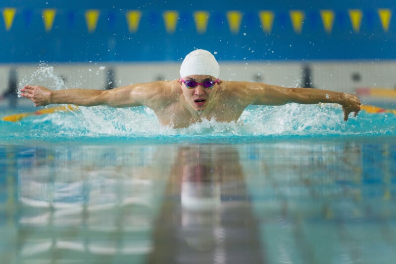 A swimmer wearing a white cap and goggles performs the butterfly stroke in a pool, with water splashing around and lane markers visible in the background.