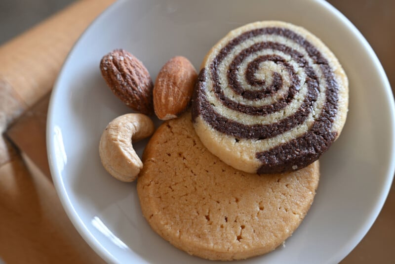 A white bowl containing two cookies—one vanilla and one chocolate-vanilla swirl—alongside two almonds and a cashew nut.