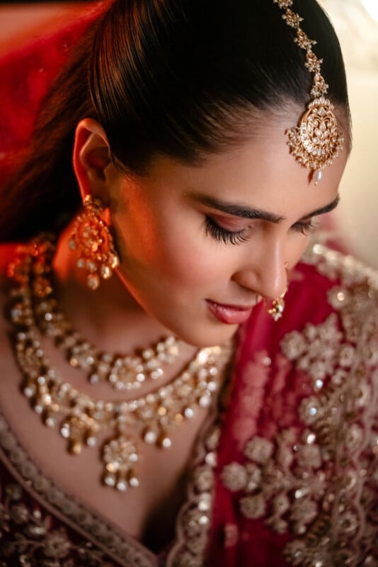 A woman dressed in traditional Indian attire with intricate gold jewelry, including earrings, a necklace, and a maang tikka, looks down gracefully. She wears a red, embellished saree and has elegant makeup.