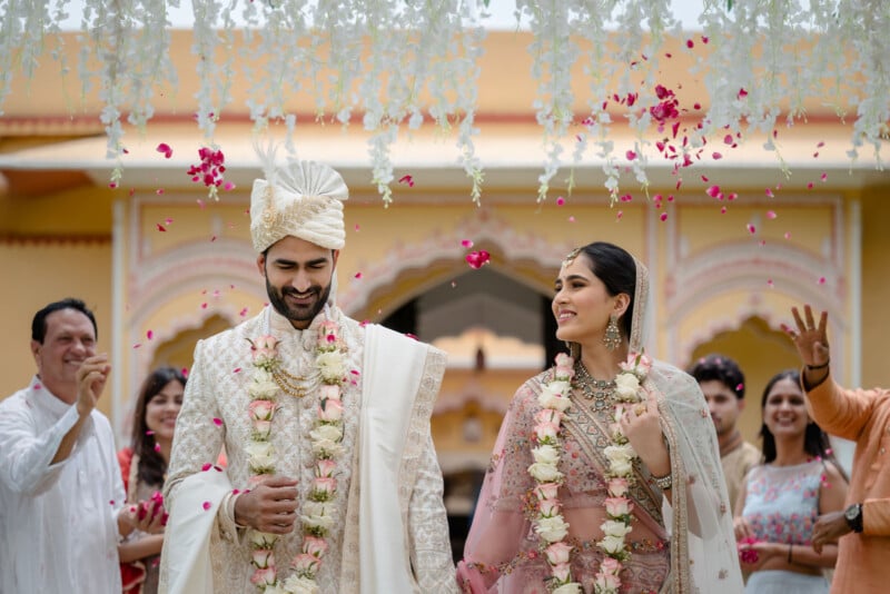 A newlywed couple in traditional Indian attire smiles as flower petals are showered on them, surrounded by joyful family and friends during their wedding ceremony under white floral decorations.
