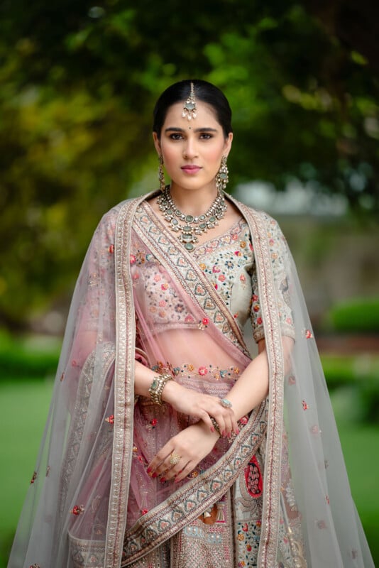 A woman wearing an ornate, pastel-colored traditional Indian lehenga with detailed embroidery, jewelry, and a sheer dupatta stands outdoors against a blurred green background.