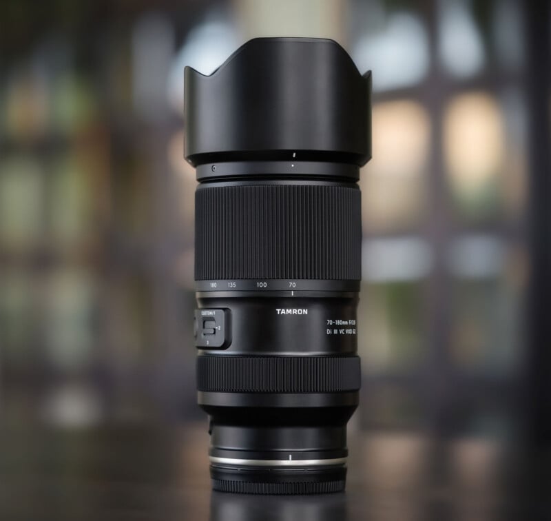 A Tamron camera lens with a lens hood is standing upright on a dark surface, with a blurred background of green and brown tones. The zoom and focus rings, as well as markings, are visible on the lens body.