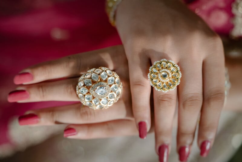 Close-up of hands with pink nail polish wearing two ornate, gold rings adorned with white gemstones, against a blurred pink and beige background.