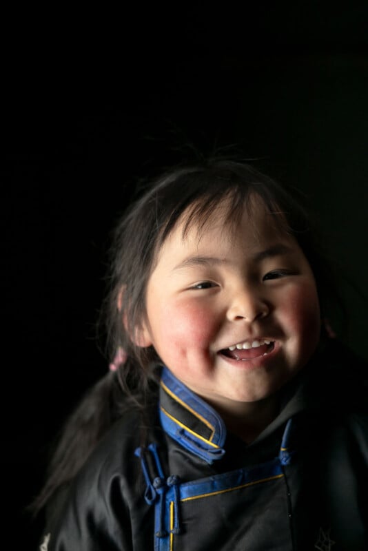 A young child with rosy cheeks and pigtails smiles brightly, wearing a dark jacket with blue and yellow trim against a black background.