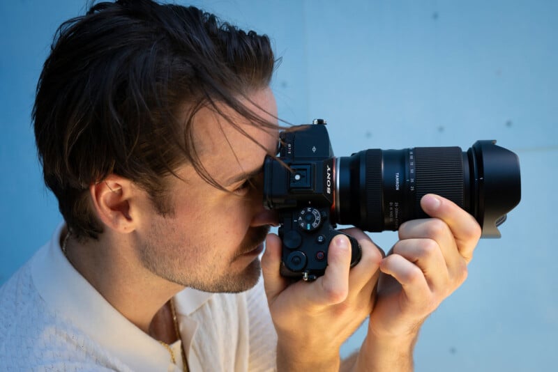 A man with brown hair and a light shirt holds a Sony camera up to his eye, focusing through the viewfinder against a light blue background.