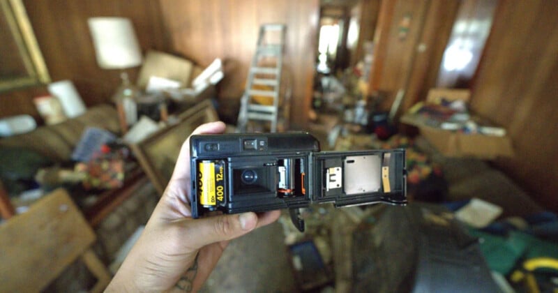 A hand holds an open 35mm film camera loaded with Kodak film; the background shows a cluttered, dimly lit room with wooden walls, a ladder, and scattered items.