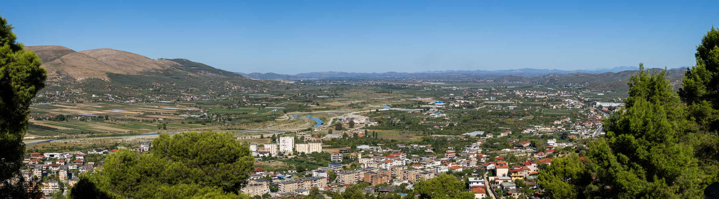 Panoramic view of a small city surrounded by greenery, hills, and mountains under a clear blue sky, with scattered buildings and a river winding through the landscape.