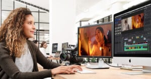 A woman sits at a desk in a bright office, editing a video on a large monitor with editing software, while others work at computers in the background. A camera and books are also on the desk.