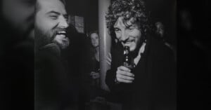 Two men laughing together at a bar, one holding a bottle, with a woman and another person in the background; black and white photo, candid and lively atmosphere.