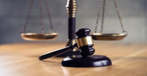 A wooden judge’s gavel rests on a sound block in front of a blurred brass balance scale, symbolizing law and justice on a wooden table.
