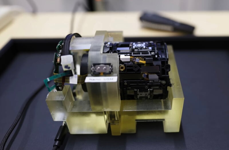 A close-up of an intricate electronic device with exposed components and circuit boards, mounted on a translucent plastic base, sitting on a dark surface. A label reads "Nano USB.