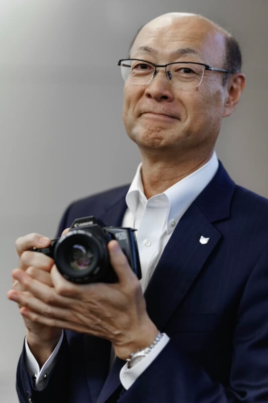 A man in a suit and glasses smiles gently while holding a camera in both hands. The background is blurred, drawing focus to the man and the camera.