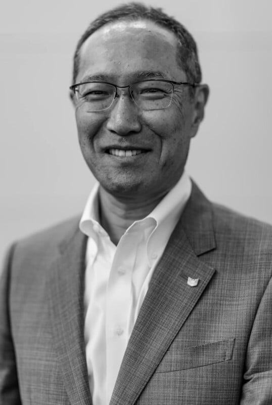 A man wearing glasses and a suit jacket over a white shirt smiles at the camera. The photo is in black and white and taken against a plain background.