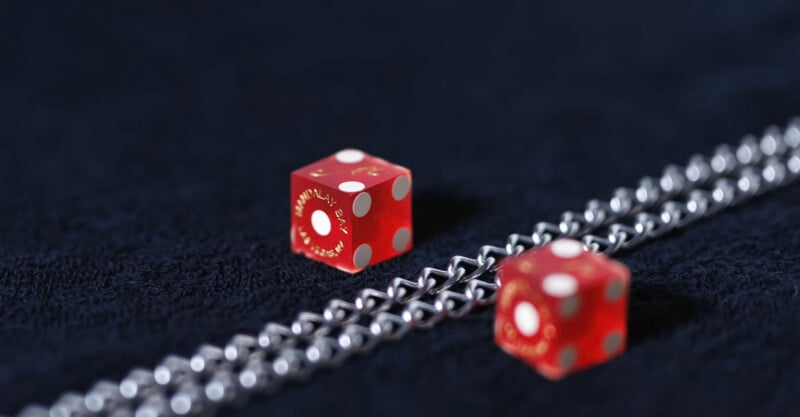 Two red dice with white dots rest on a dark textured surface next to two parallel silver chains. The dice are translucent and one is in sharp focus, while the other is blurred in the background.
