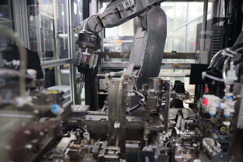 A robotic arm operates in a modern industrial setting, handling components on an automated assembly line surrounded by various machinery and metallic structures.
