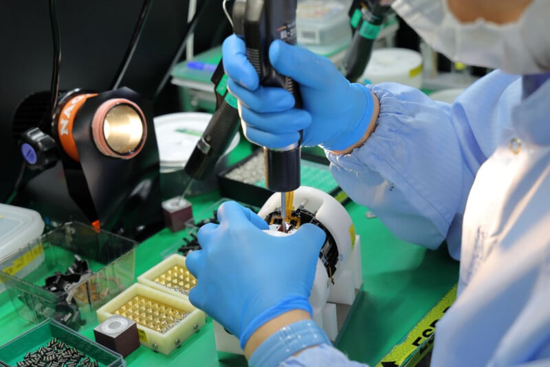 A person wearing blue gloves and a lab coat uses a power tool to assemble or repair a small electronic device at a workstation filled with components and tools.
