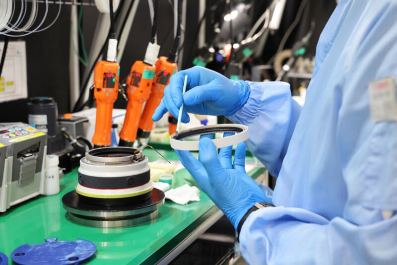 A person in blue protective gear and gloves uses a brush to clean or apply a substance to a circular component in a lab, with tools and equipment on a green workstation in the background.