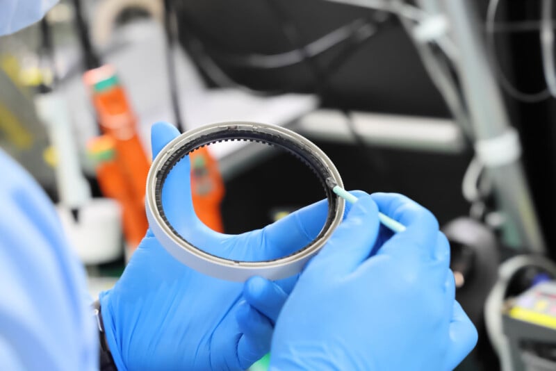 A person wearing blue gloves uses a small tool to clean or inspect a round metallic component with gear-like edges in a laboratory or technical setting.