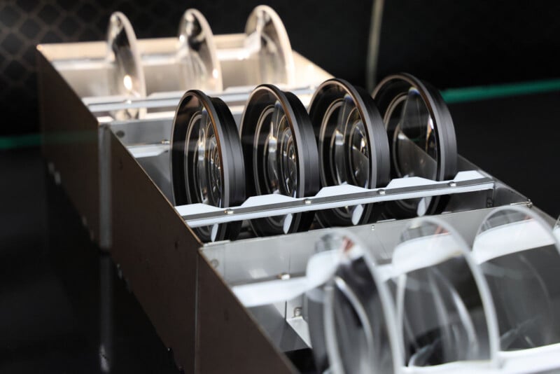 Rows of large, clear optical lenses are neatly arranged in a metal holder, each separated by dividers, against a dark, blurred background.