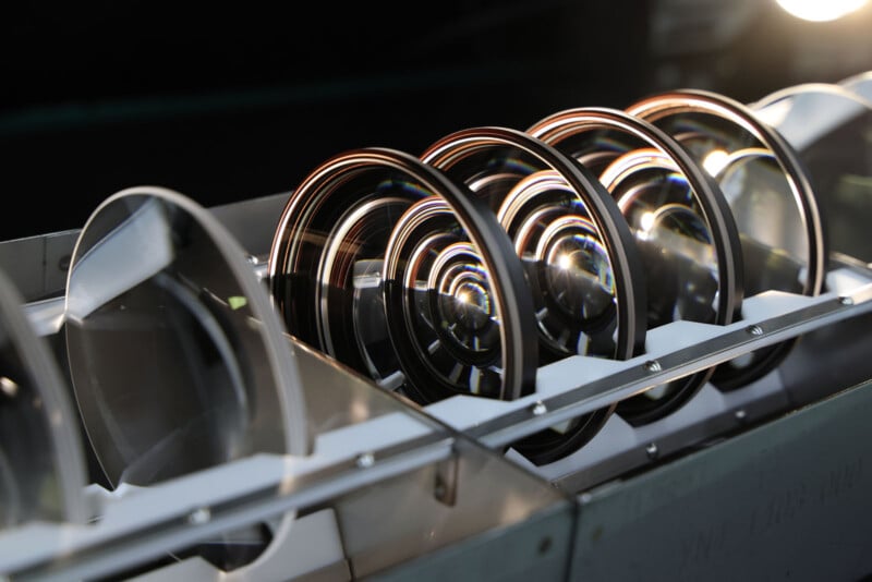 A row of large, circular glass camera lenses aligned side by side in a metal holder, reflecting light and creating curved highlights on their surfaces.