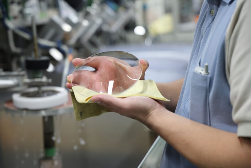 A person in a blue shirt holds a large, clear optical lens with both hands, using a yellow cloth, in what appears to be a laboratory or manufacturing setting with blurred equipment in the background.