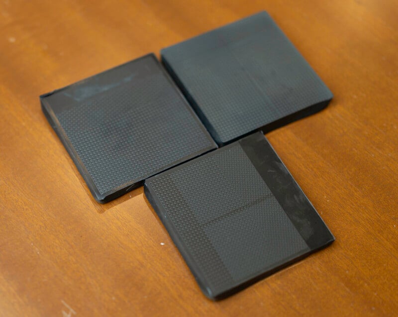Three black, square-shaped objects with textured surfaces are arranged on a wooden surface, forming an inverted "L" shape.