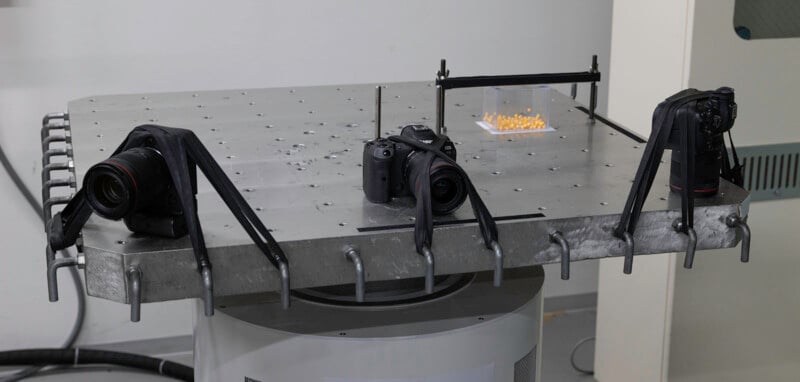 A metal platform with three cameras strapped down, facing a small group of yellow objects on a clear stand, likely set up for a scientific or technical experiment.