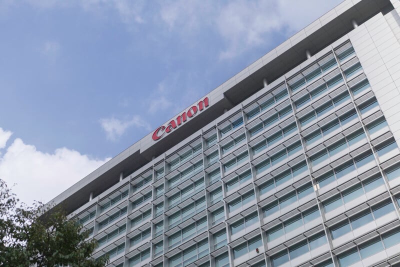 A tall, modern glass office building with the red Canon logo at the top, viewed from below against a partly cloudy blue sky. Some tree branches are visible in the lower left corner.