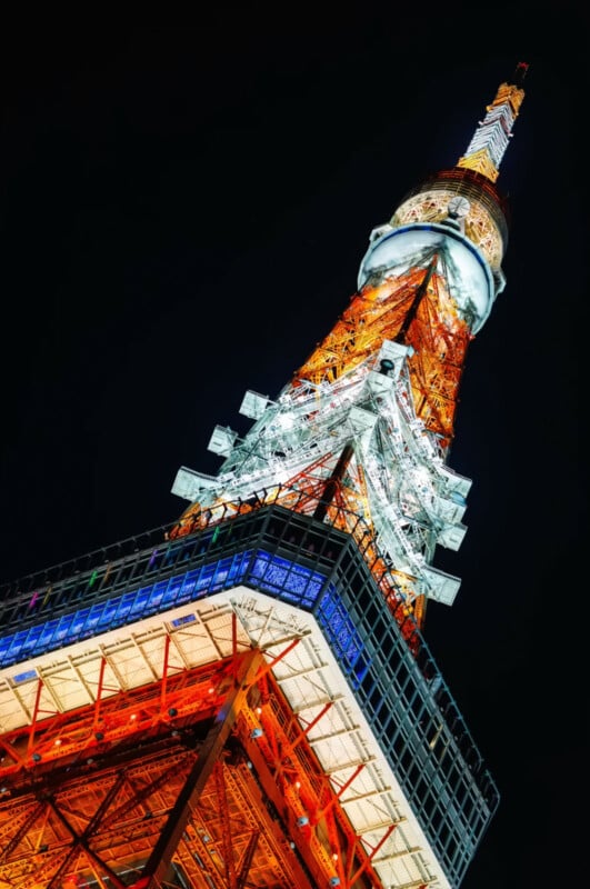 A close-up, upward view of Tokyo Tower at night, brightly illuminated with white, orange, and blue lights against a dark sky.