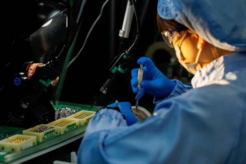 A person in protective clothing and gloves assembles electronic components at a workstation under bright lighting, using precision tools.