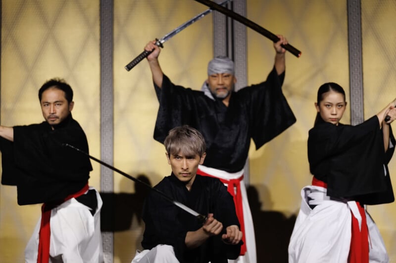 Four people in traditional black and white martial arts attire, each holding swords in dynamic poses, stand against a gold and brown patterned background, appearing focused and intense.
