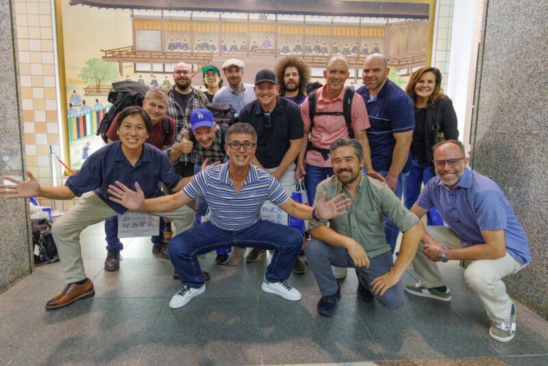 A group of fifteen smiling people, mostly men, pose together indoors in front of a large traditional mural. Four men kneel or squat at the front while the rest stand behind. Everyone looks happy and casual.