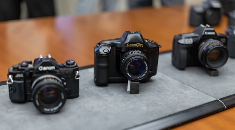 Three vintage Canon film cameras are displayed on a gray fabric mat atop a wooden table, with the center camera labeled “Canon T50.” The background is softly blurred.