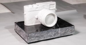 A realistic camera sculpture made of white marble sits on a rough, rectangular black stone base, displayed on a neutral surface background.